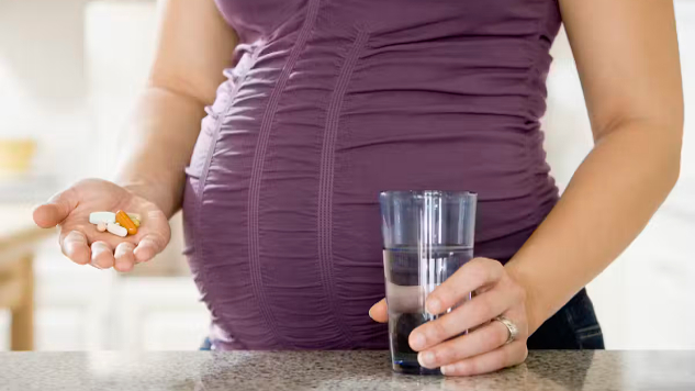 A close-up of a pregnant woman holding a glass of water in one hand and a prenatal vitamin bottle in the other